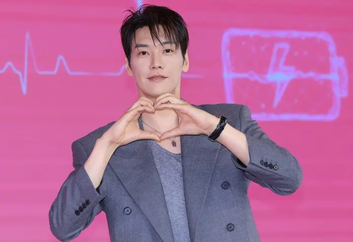 A man in a grey suit making a heart shape with his hands against a pink background featuring a heartbeat graphic.