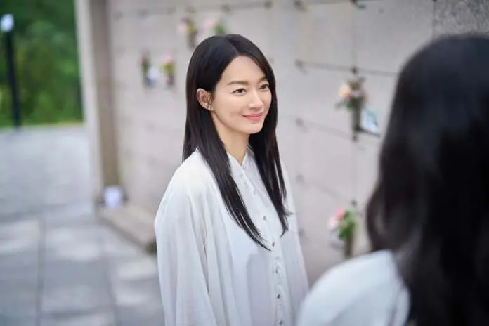 Shin Min-a wearing a white blouse smiles gently while looking into a mirror, with a stone wall adorned with flowers in the background.