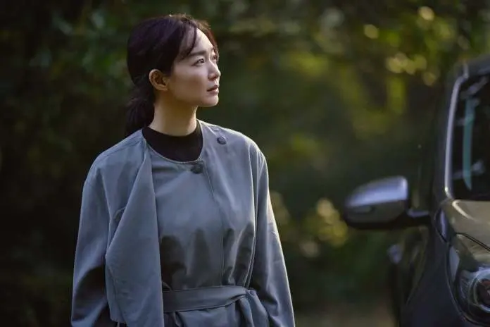 Shin Min-a in a gray outfit stands beside a car, gazing thoughtfully into the distance, with a blurred green background.
