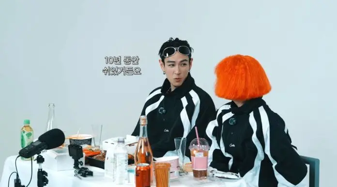 T.O.P and Nana sitting at a table with food and drinks, wearing black and white outfits. NANA has short orange hair and T.O.P has a stylish hairstyle with sunglasses.