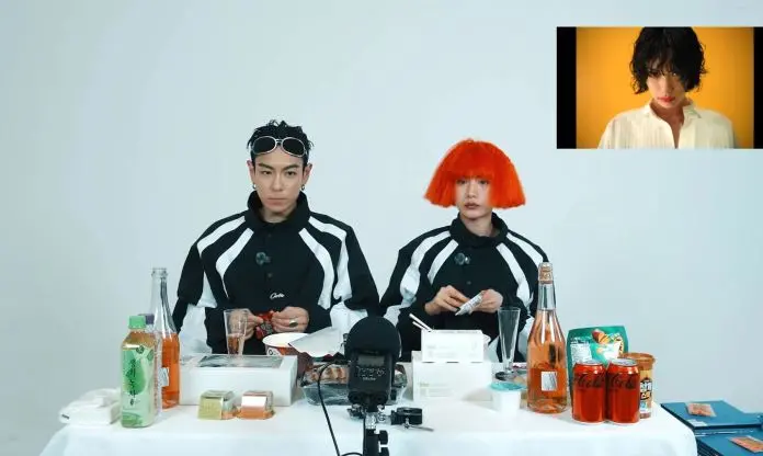 T.O.P and Nana sitting at a table set with various snacks and beverages, wearing matching black and white outfits. NANA has an orange wig and T.O.P has a serious expression. A video screen showing NANA is visible in the top right corner.