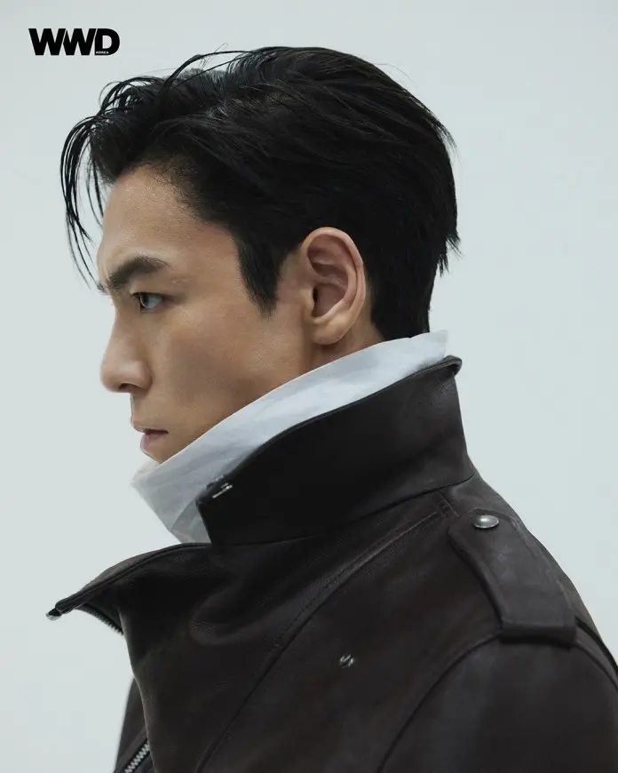 Profile view of T.O.P with dark, styled hair wearing a brown leather jacket with a high collar and a white shirt underneath, set against a neutral background.
