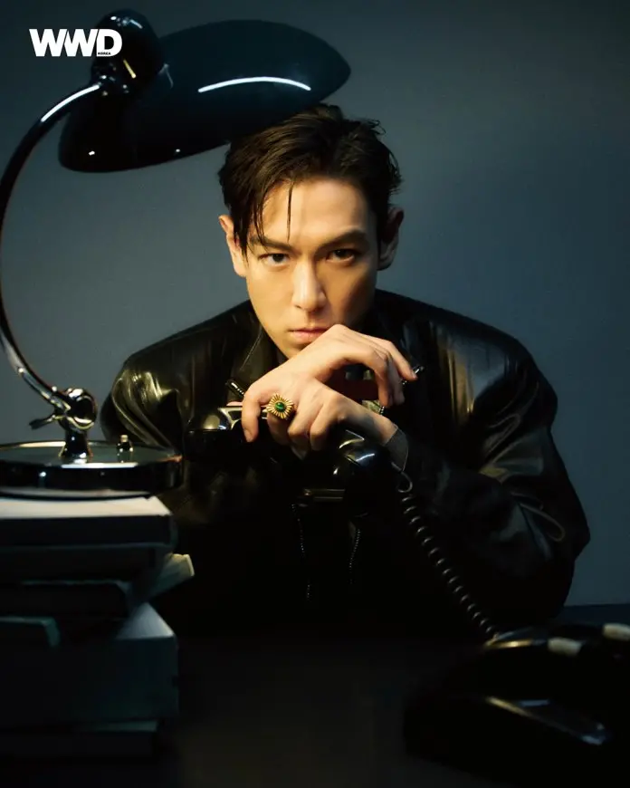 T.O.P in a black leather jacket, sitting at a desk with a telephone, looking intently towards the camera. A desk lamp illuminates the scene, highlighting his focused expression.