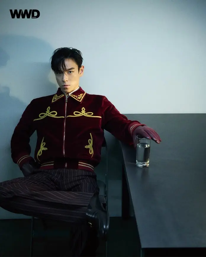 T.O.P sitting at a table wearing a red velvet jacket with gold embroidery. He has slicked back hair and is wearing gloves, holding a glass of water.