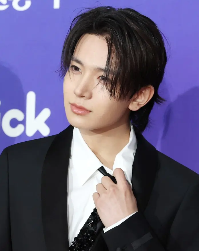 A young man in a black suit with a white shirt and black tie, adjusting his tie and looking thoughtfully at the camera, set against a purple background.