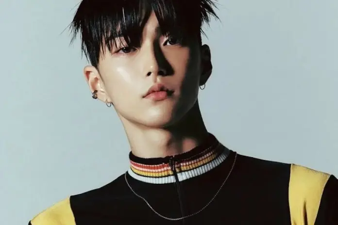 A close-up portrait of a young person with dark hair styled upwards, wearing a black outfit with colorful stripes. The background is light gray, creating a contrast with the subject's features.