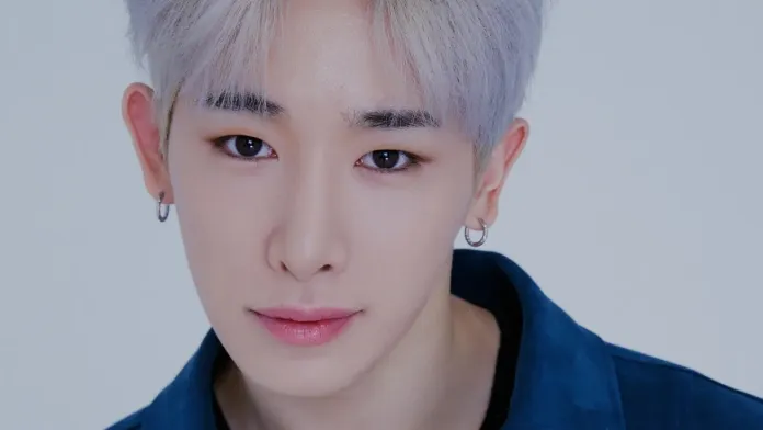 Close-up portrait of a young man with light blonde hair and hoop earrings, wearing a blue jacket and looking directly at the camera.
