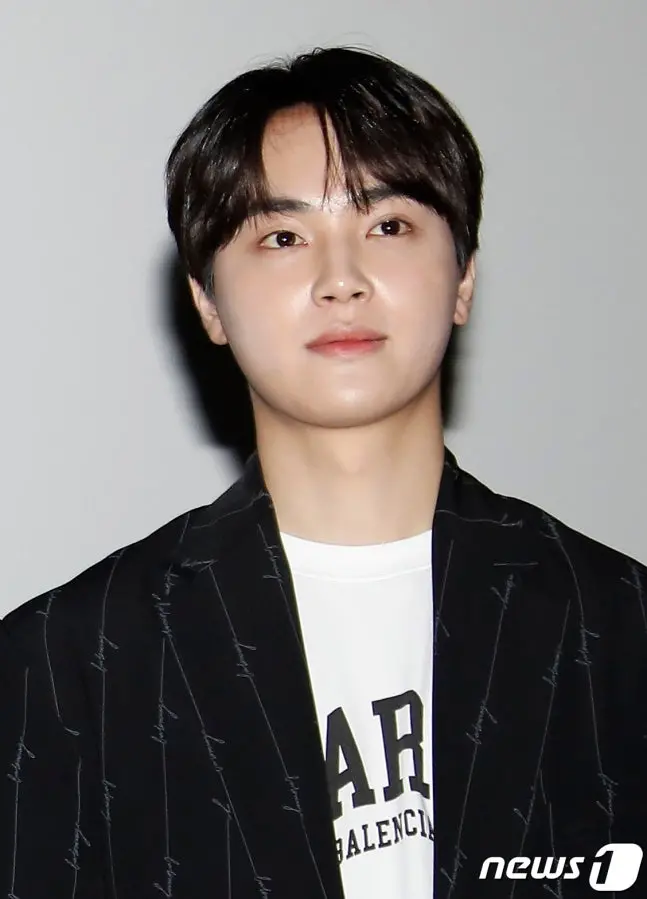 A young man with short, dark hair wearing a black pinstripe blazer over a white shirt, standing in a neutral background, looking directly at the camera.