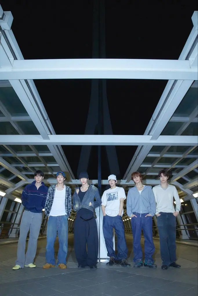 RIIZE standing on a modern bridge at night, dressed in casual clothing with a variety of styles, illuminated by soft overhead lights.