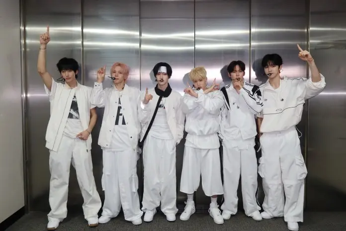 RIIZE posing together in casual white outfits, with some wearing headphones and making hand gestures, inside a well-lit indoor setting.