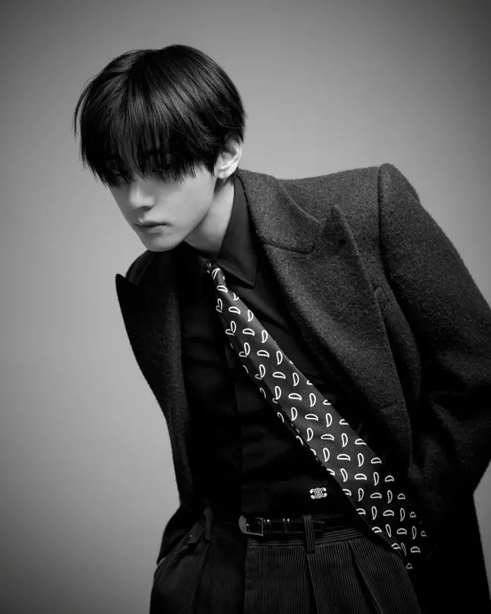 V with styled black hair, wearing a tailored coat over a black shirt and a patterned tie, gazing downwards.