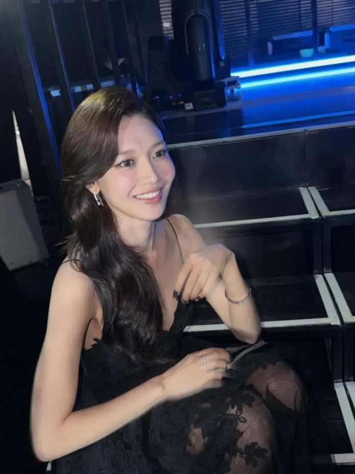 Sooyoung with long hair smiling while sitting on stairs, wearing a black dress in a dimly lit environment.
