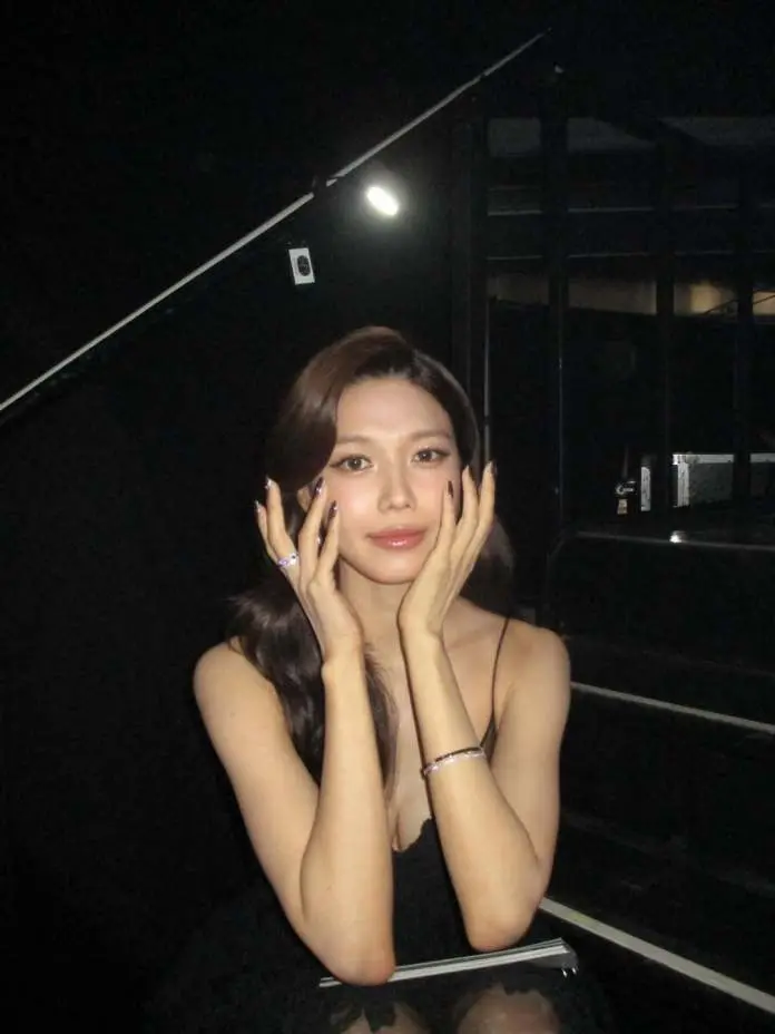 Sooyoung sitting against a dark background, gently holding her face with both hands. She has long hair and is wearing a black outfit, with visible nail polish and jewelry on her wrists.
