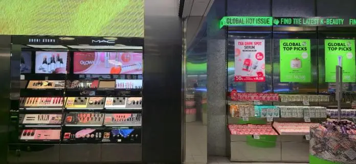 A retail display featuring cosmetics brands Bobbi Brown and MAC on the left, showcasing various makeup products including lipsticks and eyeshadows. On the right, promotional signage for K-beauty products, highlighting the 'Global Hot Issue' and 'Top Picks,' with a prominent advertisement for TXA Dark Spot Serum.
