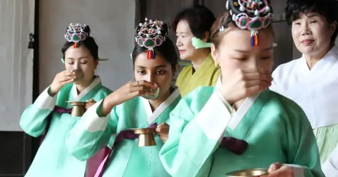 A group of women dressed in traditional green garments, participating in a tea ceremony, holding small cups and drinking, with a woman in a white outfit observing in the background.