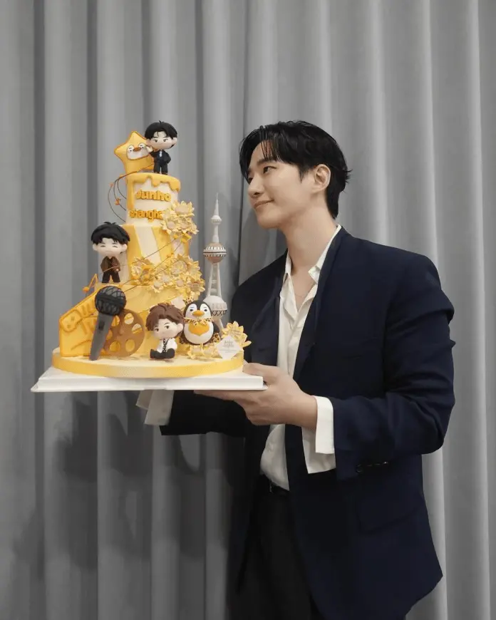 Lee Junho in a suit holding a colorful birthday cake decorated with figurines, including music and film themes, in front of a gray curtain.