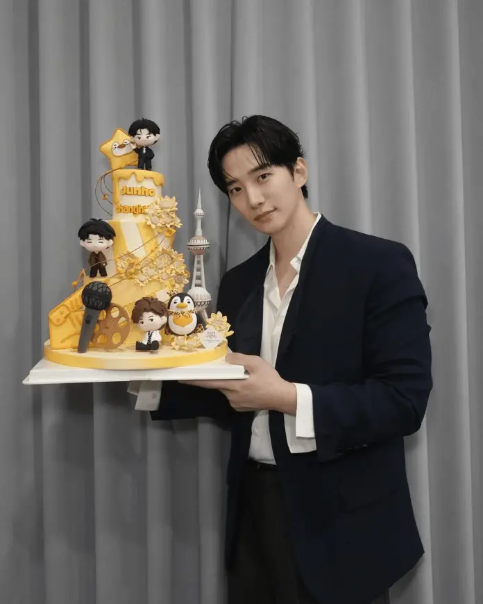 Lee Junho holding a colorful birthday cake decorated with figurines and themed elements, including a microphone and a penguin, set against a backdrop of gray curtains.
