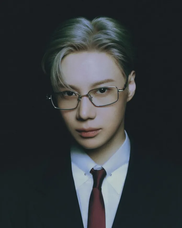Taemin with light-colored hair, wearing glasses and a suit, set against a dark background.