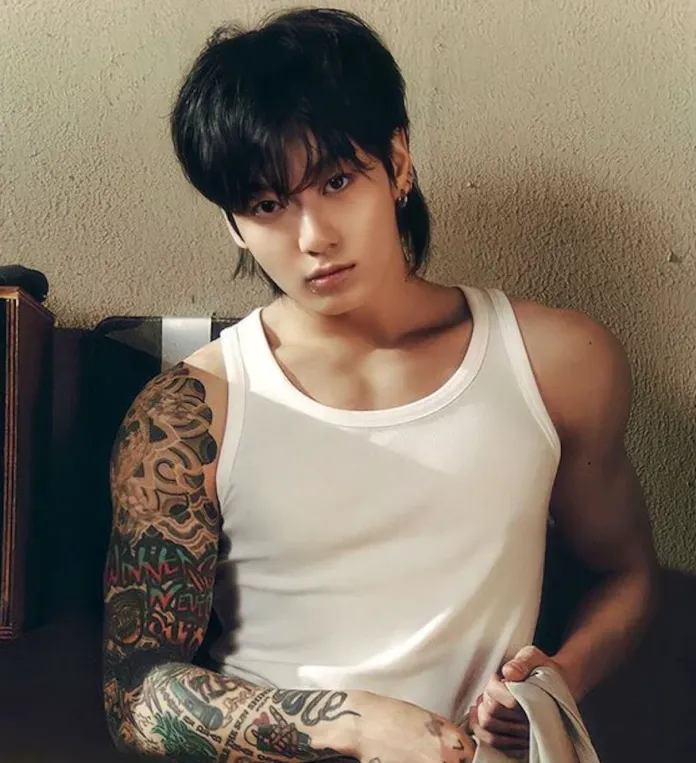 Jung Kook with long black hair and a sleeveless white shirt, showcasing intricate tattoos on their left arm, sitting against a textured wall.