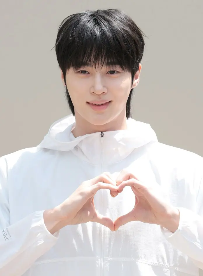 Byeon Woo-seok in a white jacket making a heart shape with their hands, smiling at the camera.
