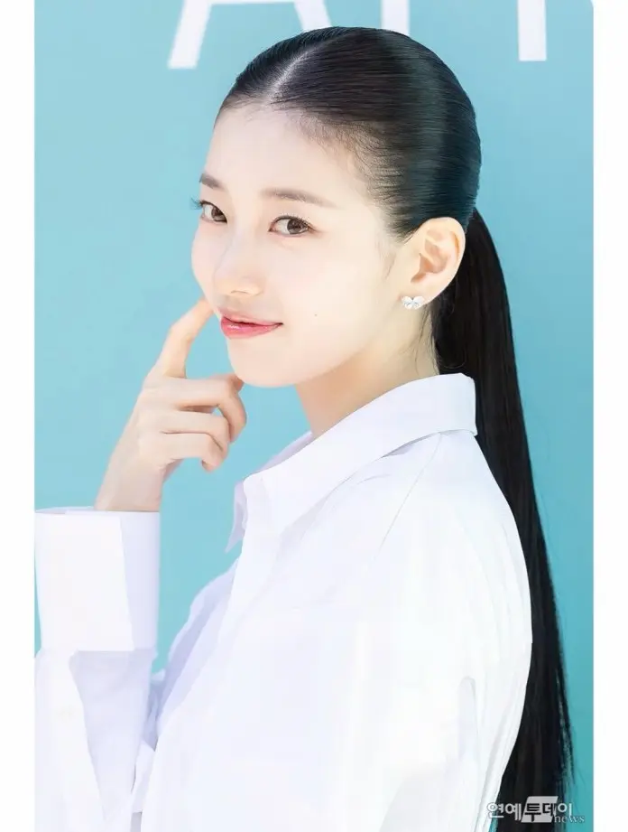 Suzy in a white shirt poses with a playful expression, touching her chin and smiling, against a light blue background.