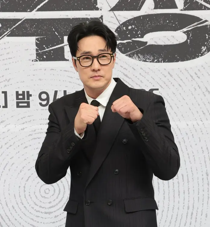 A man in a black suit and glasses poses playfully with his fists raised, showcasing a confident expression against a textured background.