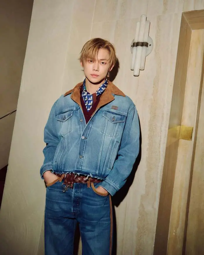 Yeonjun with blonde hair wearing a denim jacket and matching jeans, standing in an indoor setting with a light-colored wall and decorative wall light.