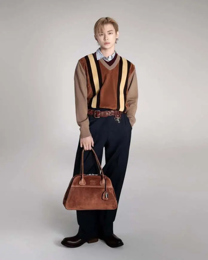 Yeonjun wearing a brown and striped sweater over a collared shirt, paired with navy trousers and brown shoes, holding a stylish brown handbag.
