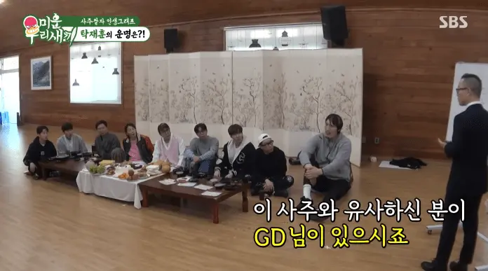 The cast of My Little Old Boy sitting on a floor with a table displaying various dishes, while a speaker stands and addresses them in a room with wooden walls and a folding screen.