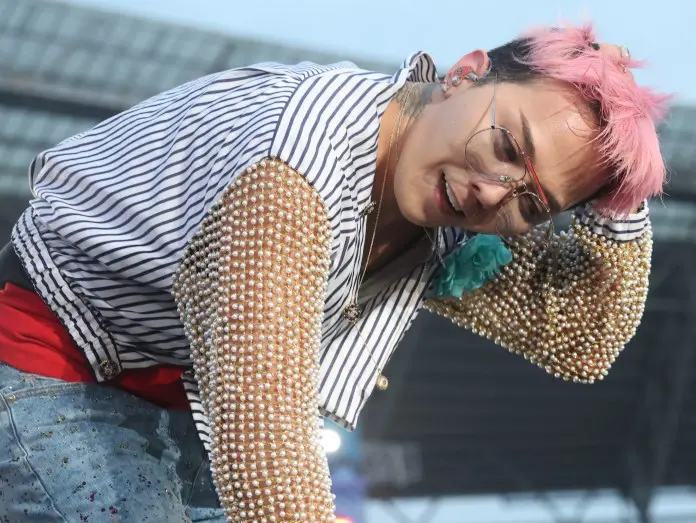 G-Dragon with pink hair and glasses wearing a striped shirt adorned with pearl embellishments, interacting with the crowd during a concert.