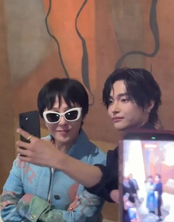 GD and Wonbin posing for a selfie, GD wearing white sunglasses and a blue jacket, and Wonbin in a black outfit, with a blurred crowd visible in the background.