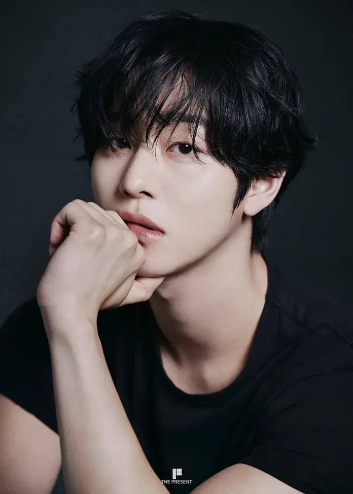 Ahn Hyo-seop with dark, tousled hair resting his chin on his hand, gazing thoughtfully towards the camera while wearing a plain black t-shirt. The background is dark, emphasizing his features.