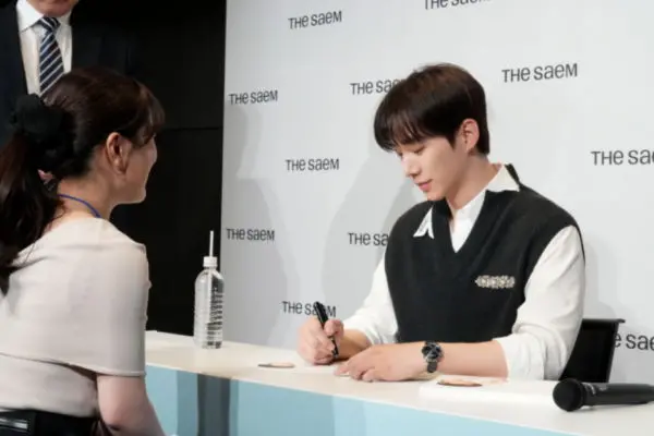 Lee Junho seated at a table, signing an autograph while facing a woman who is attentively watching him. The background features a brand logo.