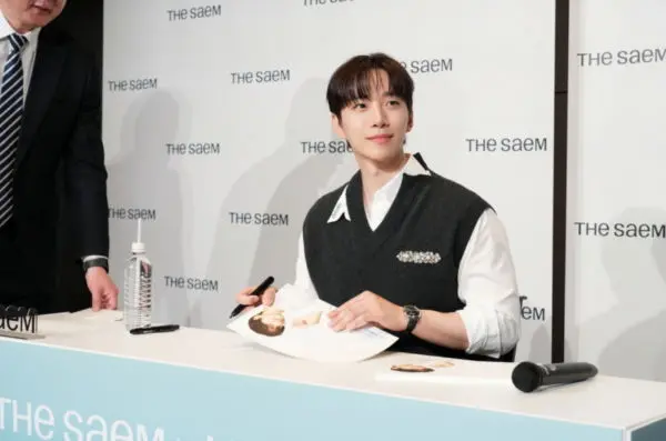 Lee Junho with styled hair smiles while sitting at a table with promotional materials for The Saem. He is wearing a sweater vest over a shirt and holding a pen, appearing ready to sign autographs.
