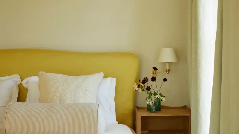 A neutral bedroom with soft butter-yellow walls, a lemon-yellow headboard, layered ivory bedding, and light neutral curtains. A small wooden nightstand with a white wall sconce and a vase of flowers.