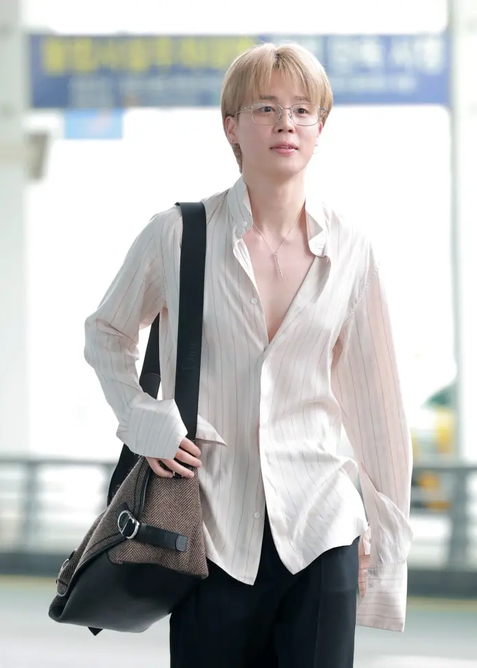 Jimin with short, light-colored hair and glasses is walking at an airport. They are wearing a striped, open-collared shirt and carrying a large bag.