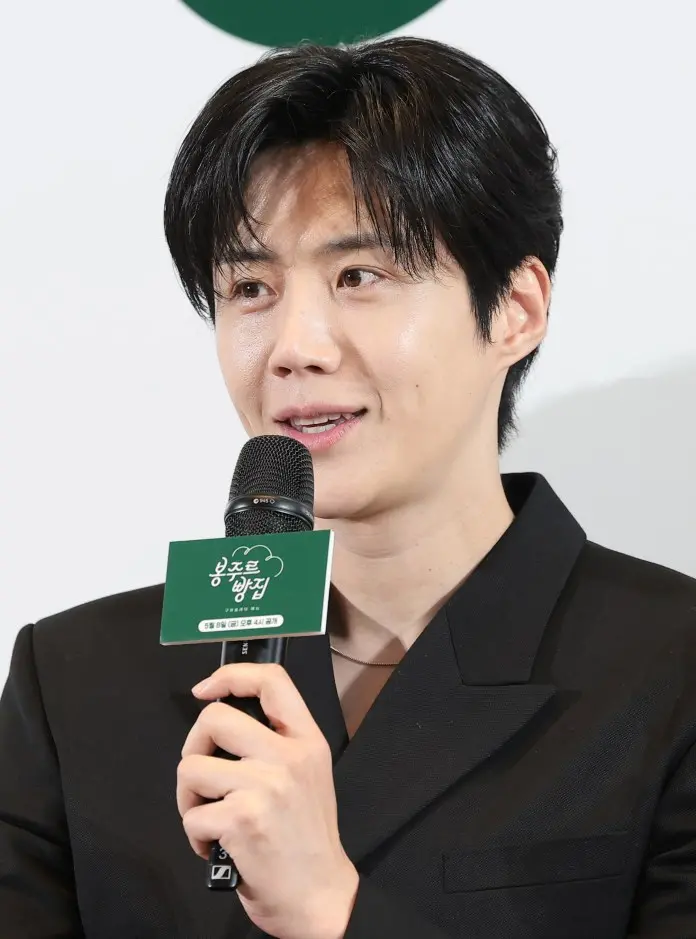 Kim Seon-ho with dark hair speaking into a microphone at an event, wearing a black blazer.