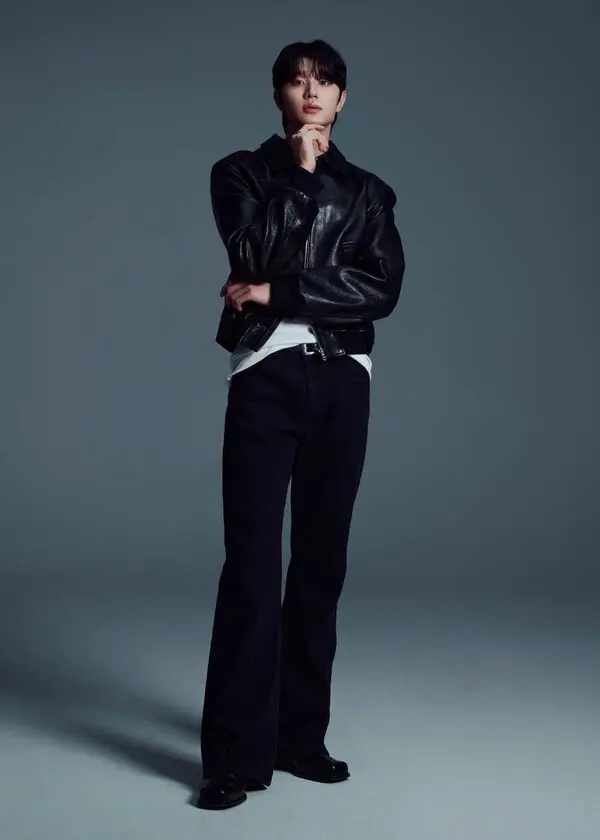 Kim Jaewon with one hand on his chin, wearing a black leather jacket, white t-shirt, and dark trousers against a gray background.