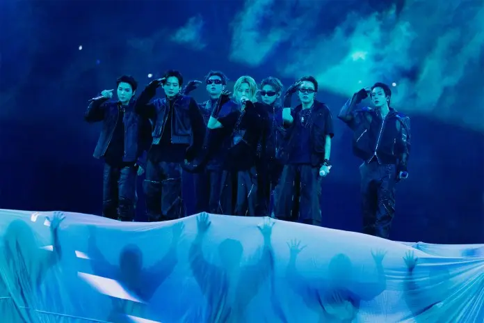 A group of seven performers on stage, dressed in black outfits, saluting while standing in front of a blue smoky backdrop, with shadowy figures visible behind a translucent curtain.