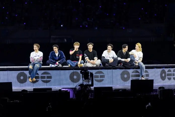 A group of eight performers sitting together on stage during a concert, with a cheering crowd in the background and stage lights illuminating them.