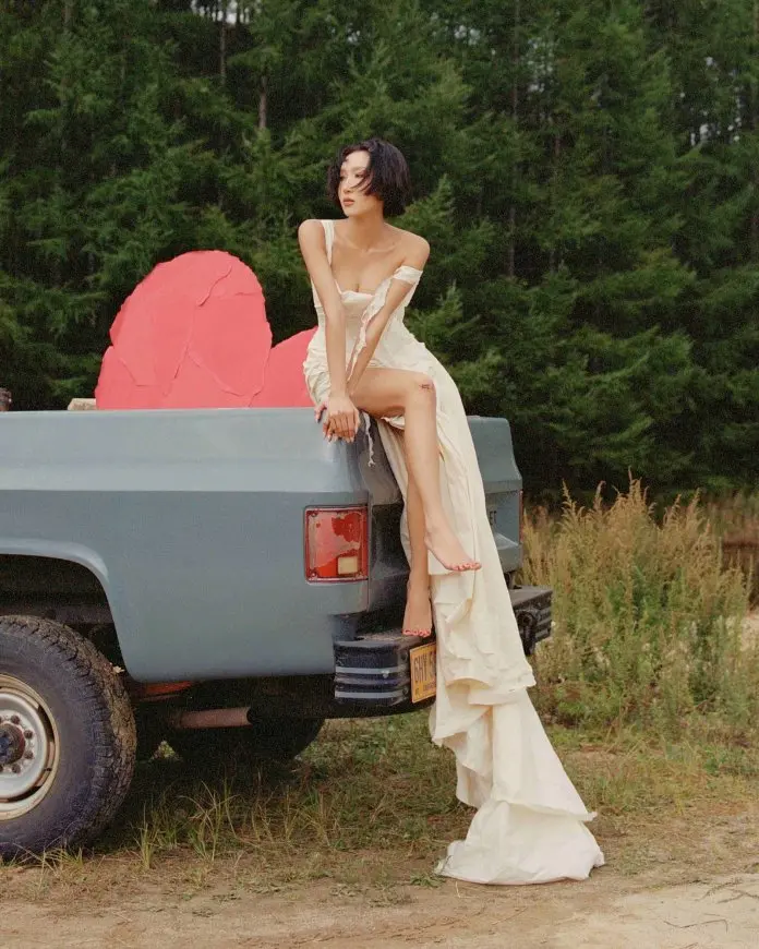 Hwasa flowing white dress poses on the back of a blue pickup truck, with a backdrop of lush green trees and a large pink rock sculpture.