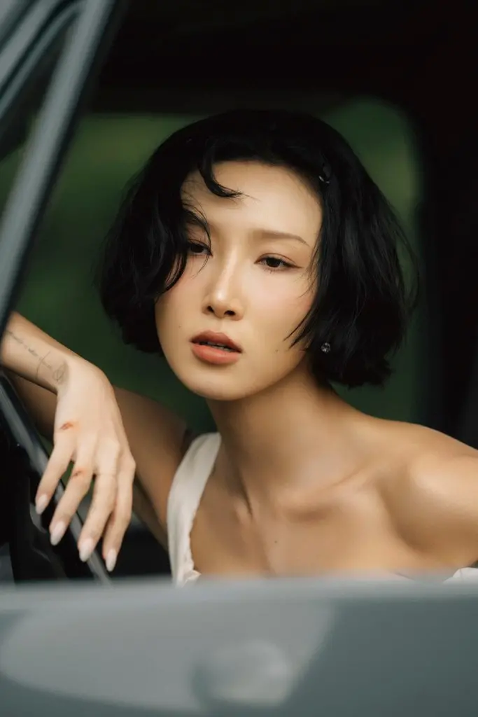 HWASA with short black hair and an elegant pose leaning out of a car window, looking contemplative with a soft expression.