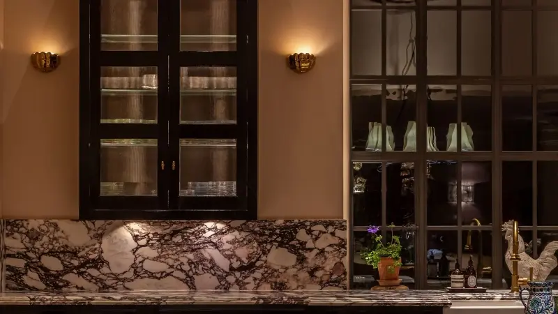 Two sconce lights are shining on the wall of a kitchen at night. There is a cabinet on the wall and red and white marble countertops.