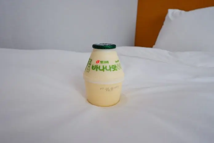 A small bottle of banana-flavored yogurt drink with a green cap, placed on a white bedspread.