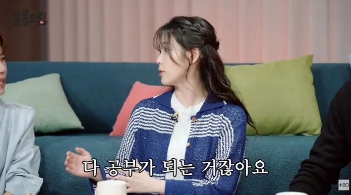 IU in a blue and white sweater is engaged in conversation with another person off-screen, sitting on a green couch. She is animatedly gesturing while speaking.