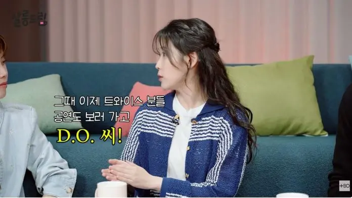 A conversation scene featuring IU seated on a couch. IU is dressed in a blue and white striped cardigan, is speaking.  Various colorful cushions are visible in the background.