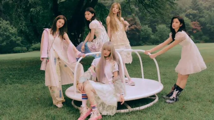A group of five young women in pastel-colored dresses posing on a circular platform in a grassy outdoor setting, surrounded by trees.