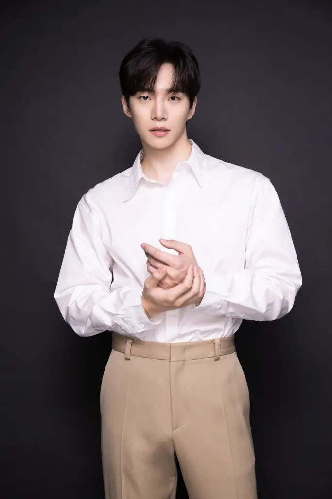 Lee Junho wearing a white shirt and beige pants stands against a dark background, with a serious expression and hands clasped together.