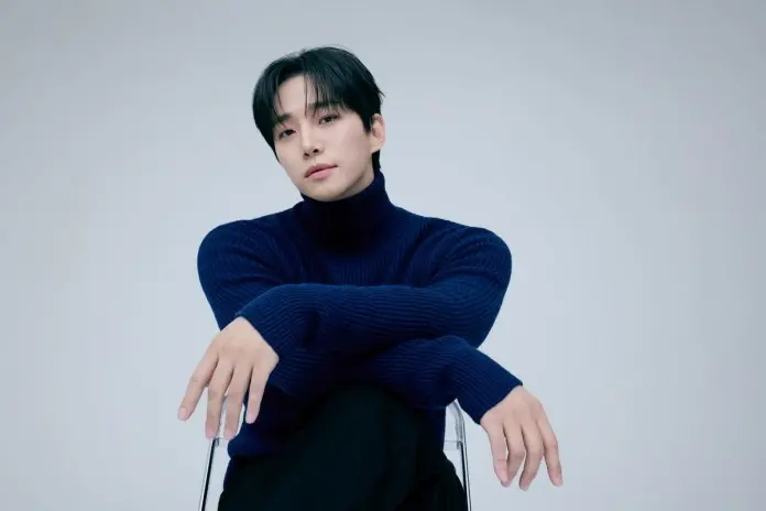 Lee Junho in a dark blue turtleneck sweater poses with his arms crossed while sitting on a clear chair against a light grey background.