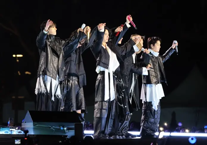 BTS dressed in black outfits on stage, posing with their arms raised, during a concert at night.